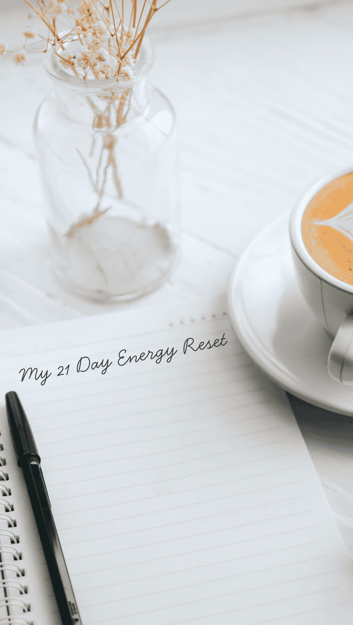 A notebook with the words My 21 Day Energy Reset written on it, a black pen, a cup of coffee, and a glass vase with dried flowers on a white table—perfect for balancing your chakras and setting mindful intentions.