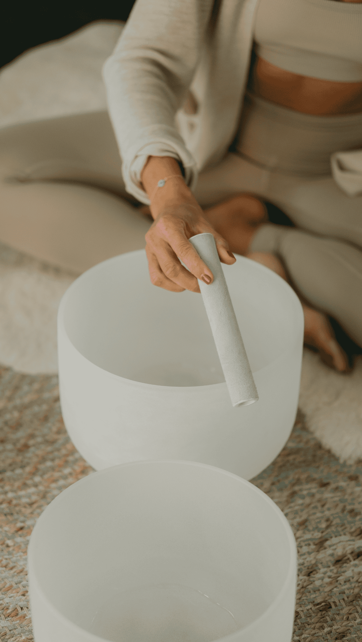 A person in beige clothing sits cross-legged on a rug, holding a white mallet over a frosted crystal singing bowl, possibly preparing for a sound healing or meditation session to balance their chakras.