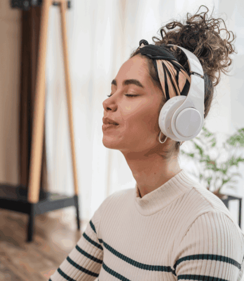 A woman with curly hair and a headband sits indoors with her eyes closed, wearing white headphones and a striped sweater, appearing relaxed and content as soothing music calms her nervous system. Light filters through the window behind her.