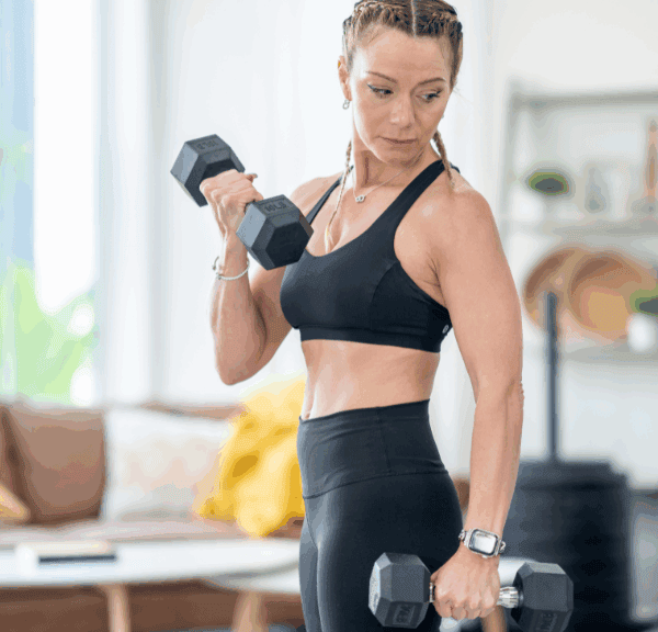 A woman in athletic wear lifts dumbbells in her bright living room, focusing on her workout. She stands near a sofa with yellow cushions, turning her space into a stylish home gym with inviting home decor in the background.