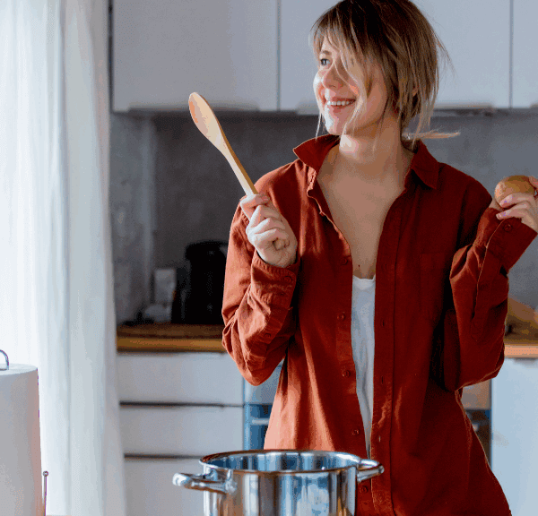 A woman in a brown shirt stands in a kitchen, smiling while holding a wooden spoon and a potato, ready for some healthy eating and meal prep with a pot on the stove in front of her.