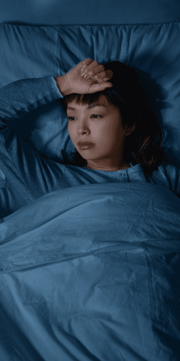 A woman lies in bed under a blue blanket, looking thoughtful or worried, with her hand resting on her forehead in a dimly lit room, perhaps contemplating sleep solutions.