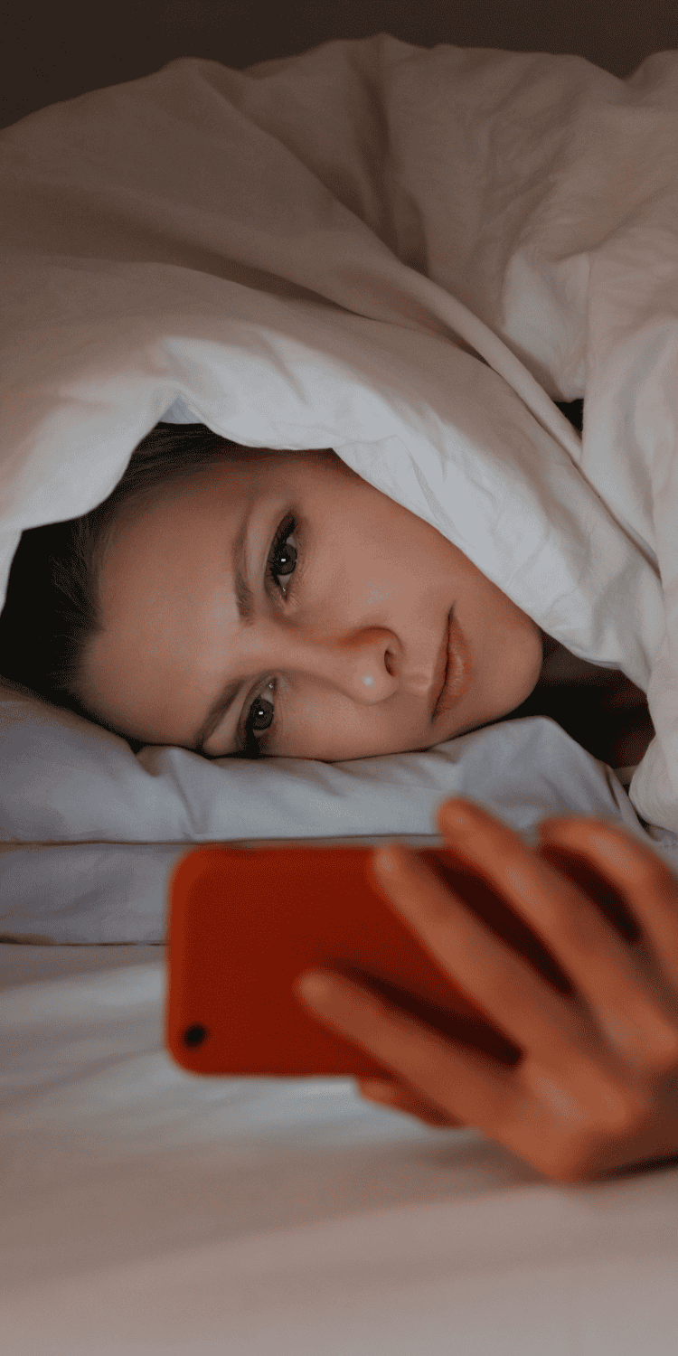 A person lying in bed under a white blanket looks at a smartphone, holding it close to their face in a dimly lit room—possibly searching for sleep solutions.
