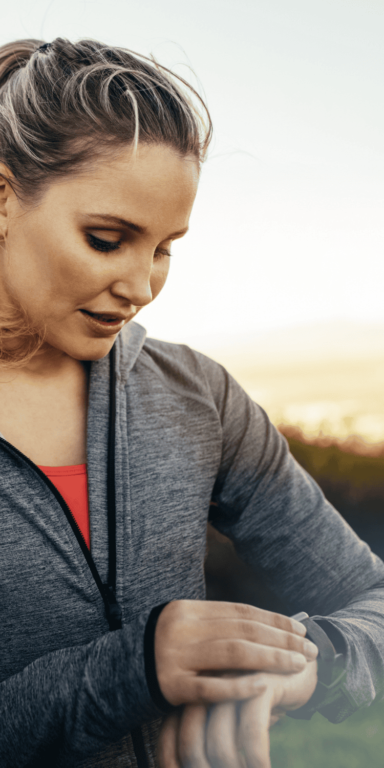 A woman in athletic wear outdoors looks down at her smartwatch, possibly checking her fitness data or exploring bio-hacking trends, with a blurred background of nature.