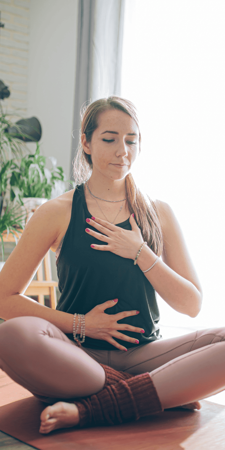 A woman sits cross-legged on a mat indoors, eyes closed, one hand on her chest and the other on her abdomen, practicing deep breathing or meditation as part of her bio-hacking routine. Sunlight streams in through a window behind her.