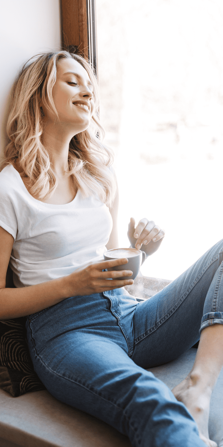 A young woman with blonde hair sits by a window, smiling with her eyes closed. She holds a coffee mug in one hand, embracing a moment of bio-hacking relaxation in her white t-shirt and blue jeans, appearing relaxed and content.