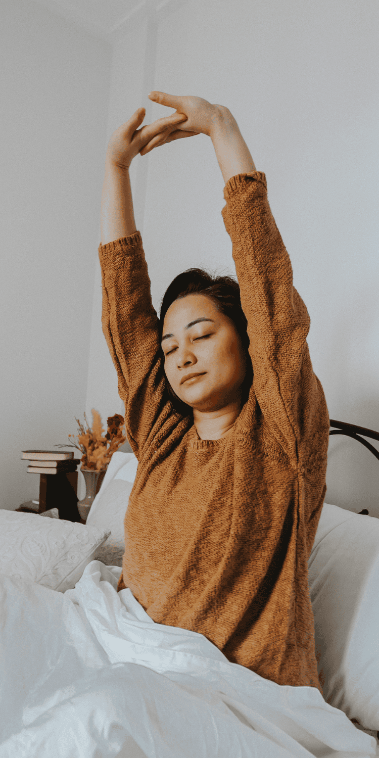 A person with medium-length dark hair sits up in bed, eyes closed, wearing a brown sweater, and stretches their arms overhead—a serene moment reflecting thoughtful Sleep Solutions. A small stack of books and dried flowers rest on the bedside table.