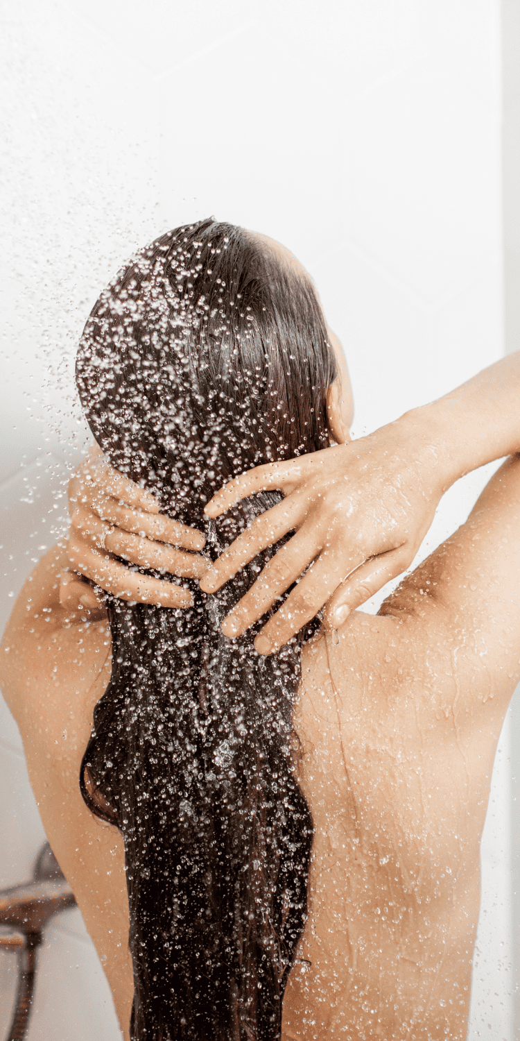 A person with long wet hair stands under a shower, facing away, hands on the back of their head as water sprays over them—a moment of bio-hacking self-care and rejuvenation.