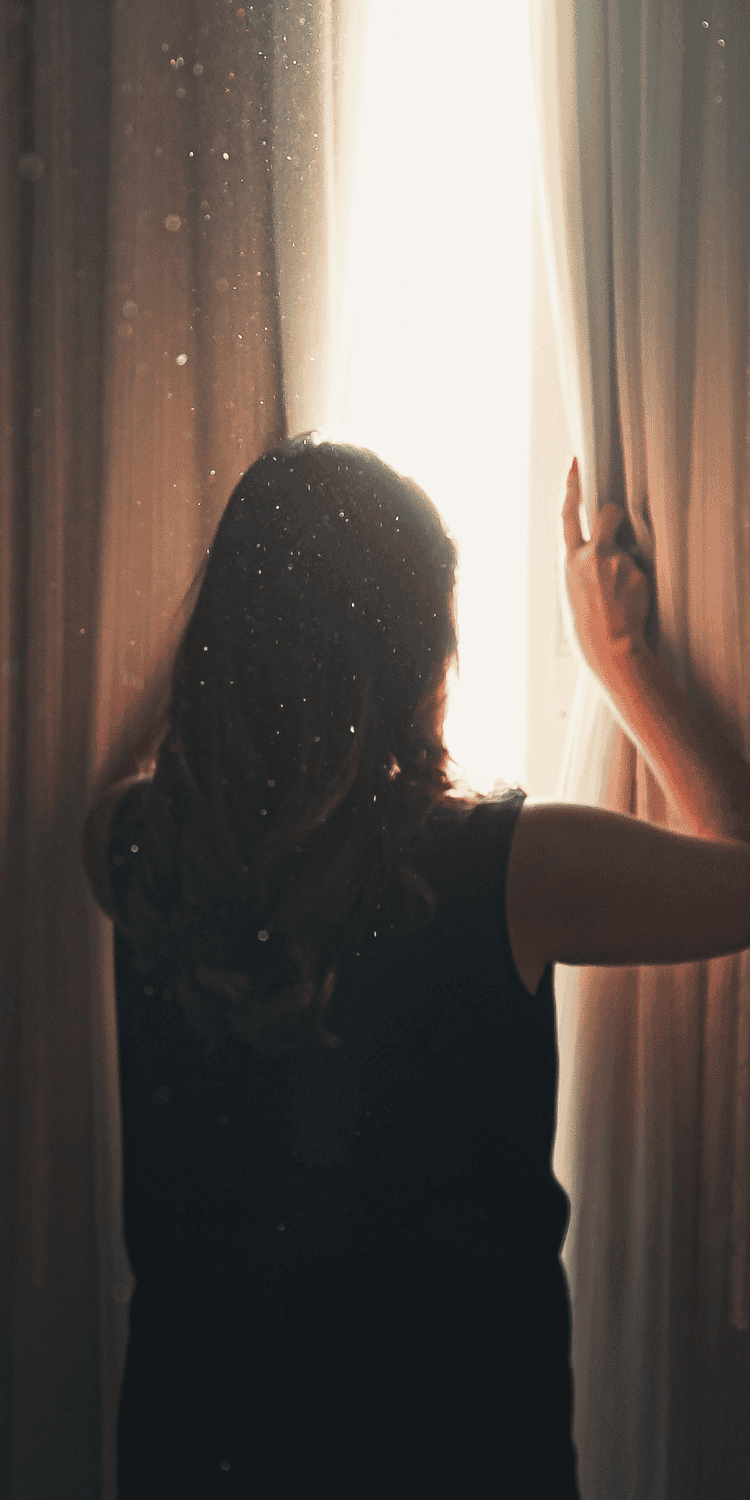 A person with long hair, seen from behind, opens beige curtains to let sunlight into a dimly lit room—an inviting moment that hints at gentle Sleep Solutions as dust particles float in the air, illuminated by the bright window light.