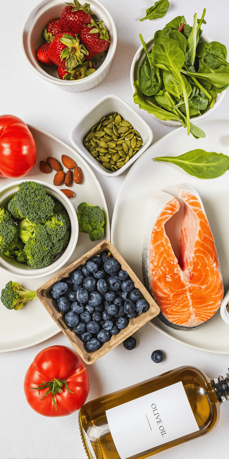 A variety of healthy foods on a white surface, ideal for bio-hacking, including fresh salmon, tomatoes, blueberries, broccoli, spinach, strawberries, almonds, pumpkin seeds, and a bottle labeled olive oil.