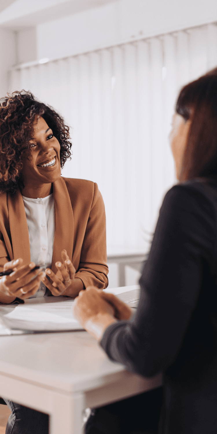 Two women sit across from each other at a desk, engaged in a conversation about Sleep Solutions. One woman is smiling and gesturing with a pen, while the other listens attentively. Bright, natural light fills the room.
