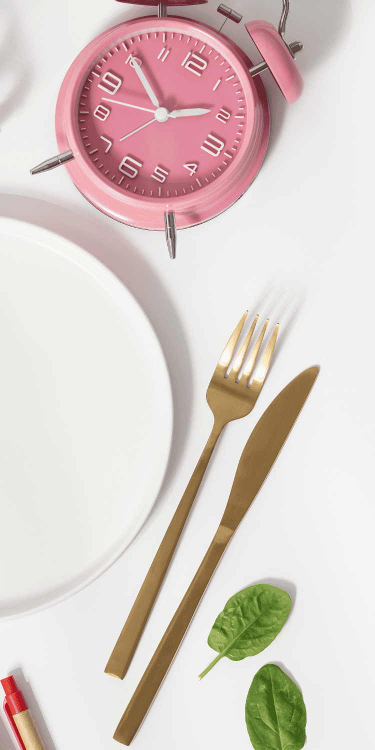 Flat lay of a pink alarm clock, a white plate, gold fork and knife, and three spinach leaves on a white background, capturing the essence of bio-hacking mealtime routines.