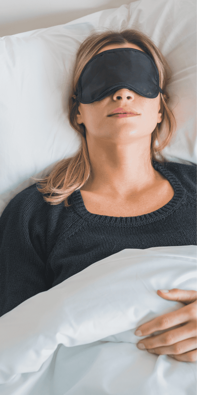 A woman practicing bio-hacking wears a black sleep mask as she lies in bed under a white blanket, resting her head on a white pillow with her hand on top of the blanket.