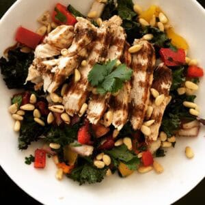 A Chicken Kale Salad featuring grilled sliced chicken, mixed greens and kale, red and yellow bell peppers, onions, pine nuts, balsamic glaze, and a cilantro garnish served in a white bowl.