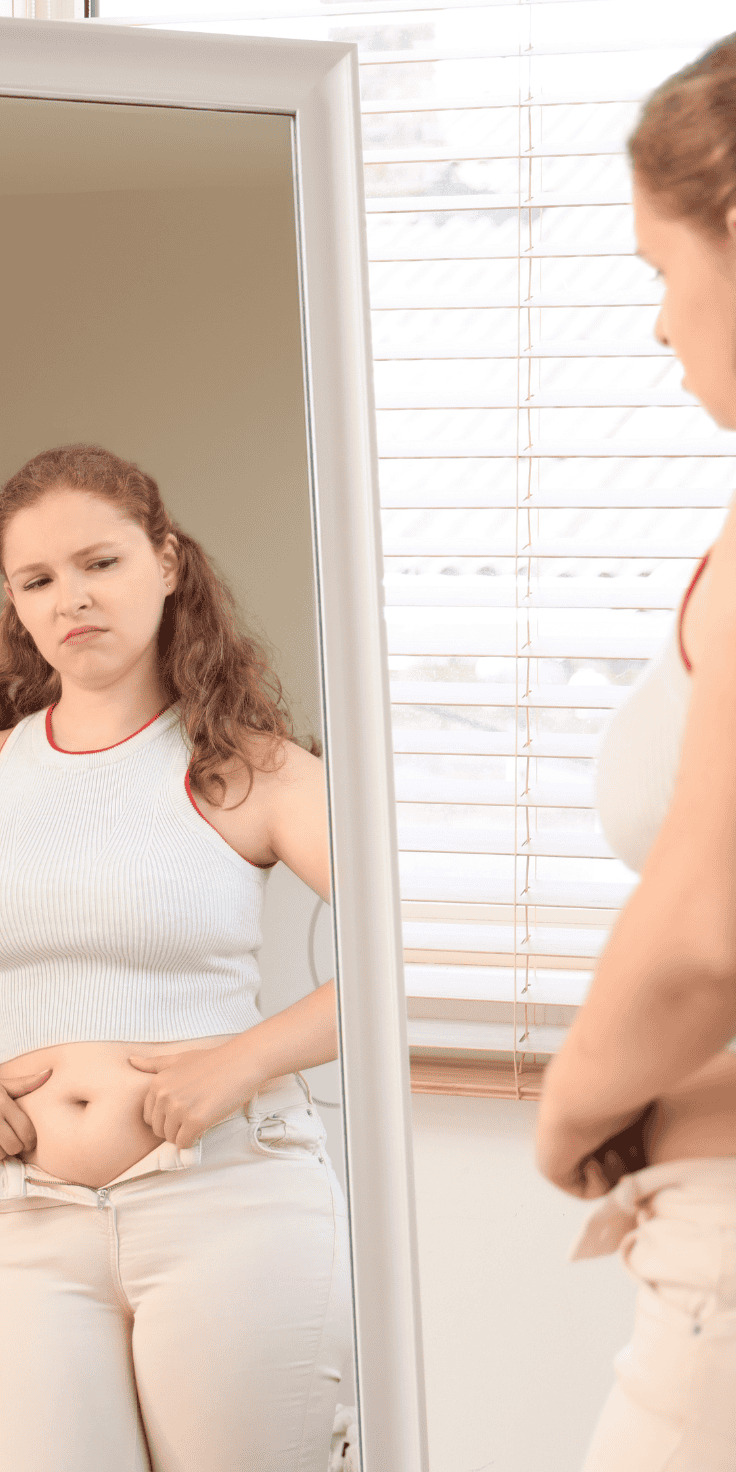 A woman stands in front of a mirror, looking at her reflection with a concerned expression, pinching the skin on her stomach as she considers starting a new diet plan. She appears to be self-conscious about her body.