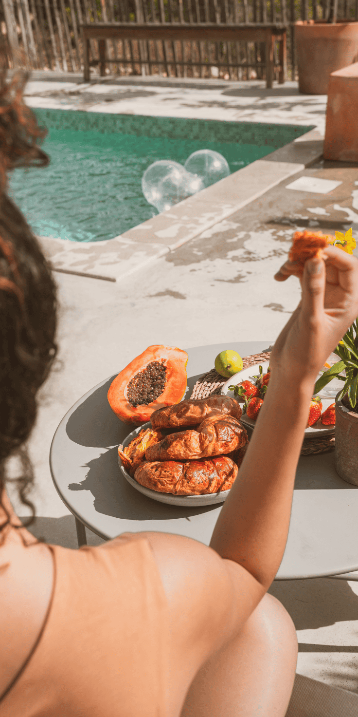 A person sits by a pool, savoring a pastry near a table with croissants, papaya, fresh fruit, and flowers—enjoying a sunny outdoor meal that feels like the perfect break from any strict diet plan.