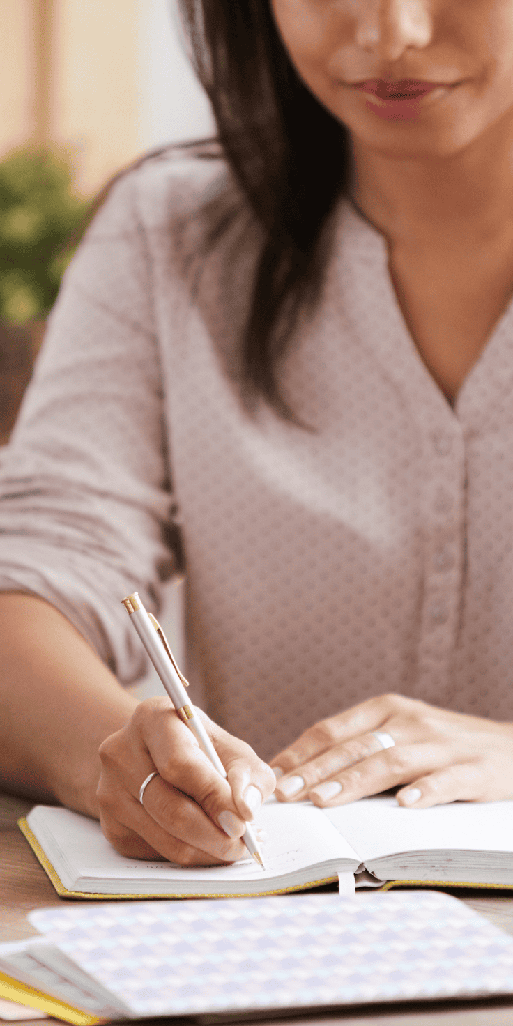 A woman wearing a light pink blouse is sitting at a table, focused on writing her diet plan in a notebook with a pen. Her face is partially visible as she concentrates on her work.