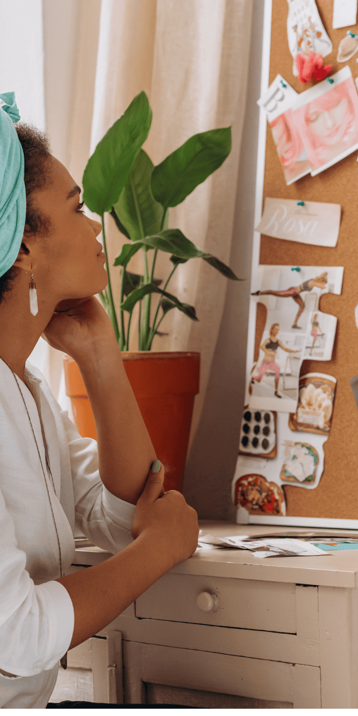 A woman sits at a white desk, thoughtfully reviewing a diet plan pinned to a corkboard filled with colorful images and notes. A potted plant and beige curtains are in the background.