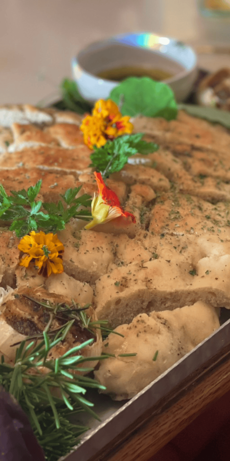 A tray of freshly baked focaccia bread from our favorite focaccia recipe is garnished with herbs, rosemary, and edible flowers, with a small bowl of olive oil in the background.