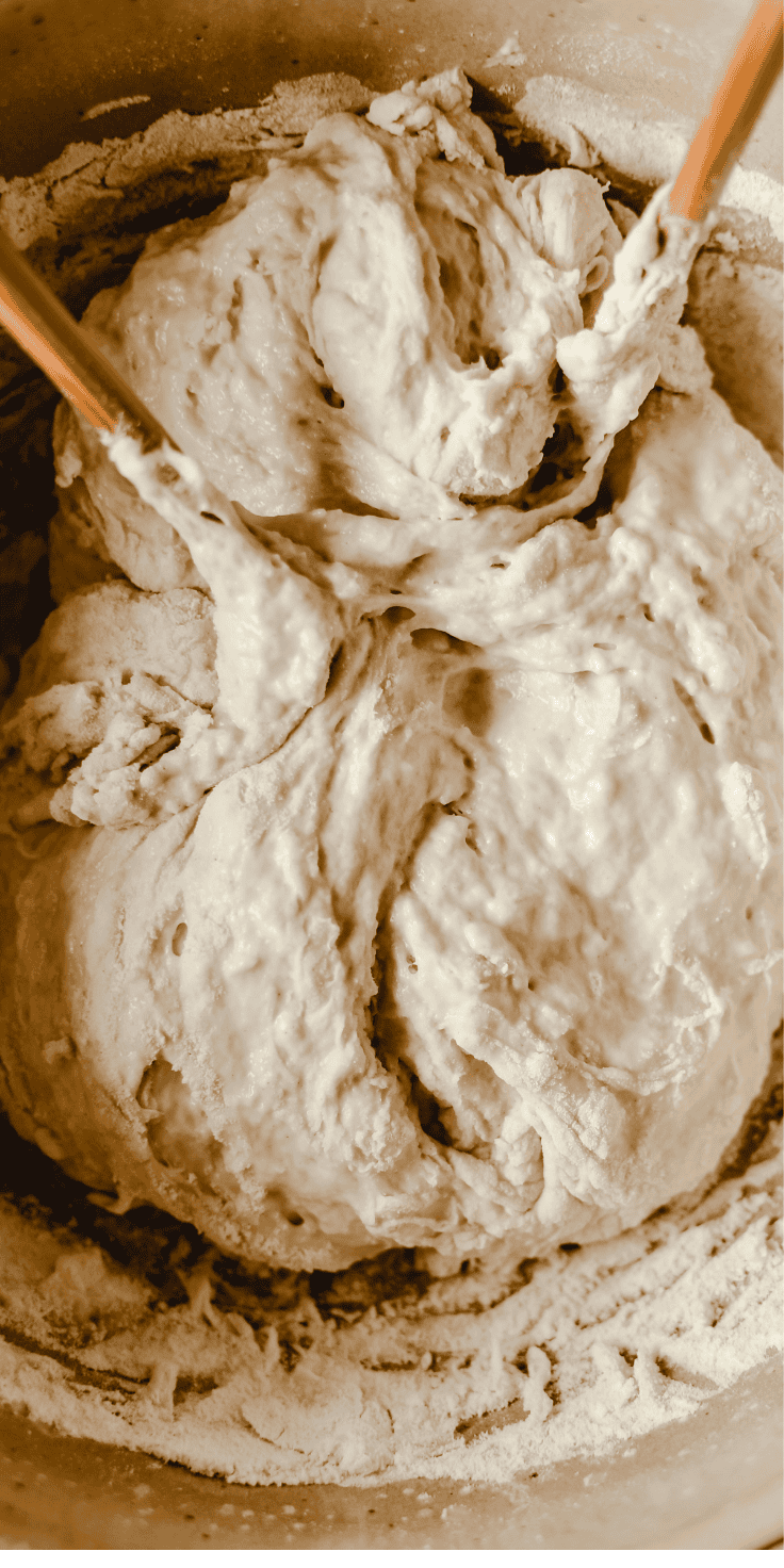 Close-up of bread dough being mixed in a bowl with wooden spoons, surrounded by flour—perfect for starting your favorite focaccia recipe.