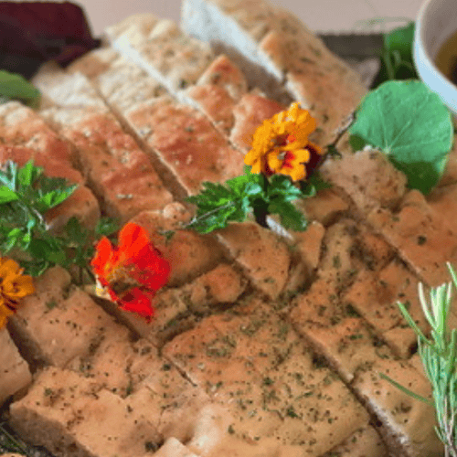 A platter of sliced focaccia bread, made from a classic focaccia recipe, garnished with herbs and edible flowers, served with olive oil and surrounded by fresh rosemary and roasted garlic.