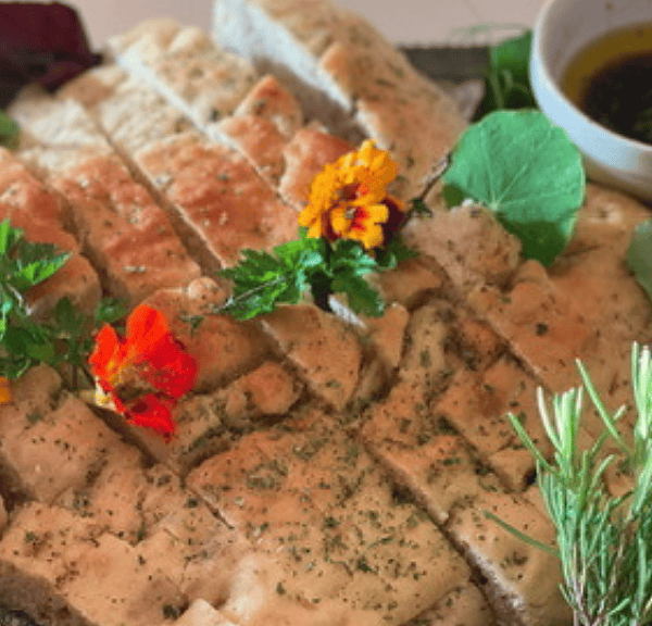 A platter of sliced focaccia bread, made from a classic focaccia recipe, garnished with herbs and edible flowers, served with olive oil and surrounded by fresh rosemary and roasted garlic.