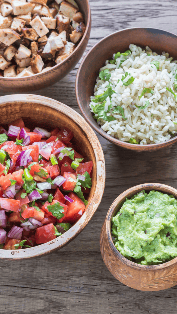 Four wooden bowls on a rustic table, each part of a vibrant Mexican Salad Bowl: grilled tofu cubes, cilantro rice, chopped tomato salsa with onion and cilantro, and creamy guacamole.