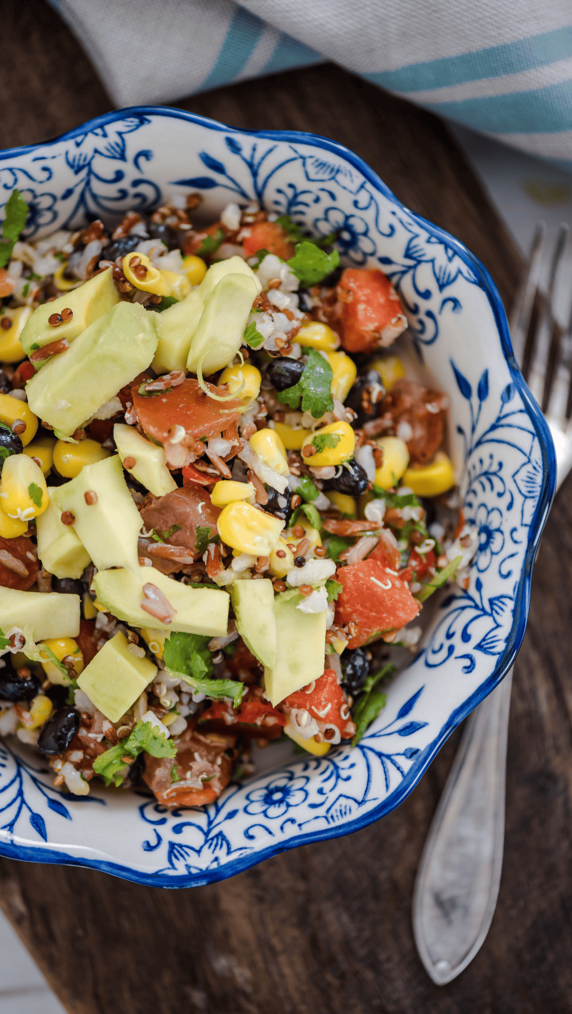 A vibrant Mexican Salad Bowl in a blue and white floral bowl, featuring avocado slices, corn, black beans, tomato, rice, and fresh herbs on a wooden surface with a fork nearby.