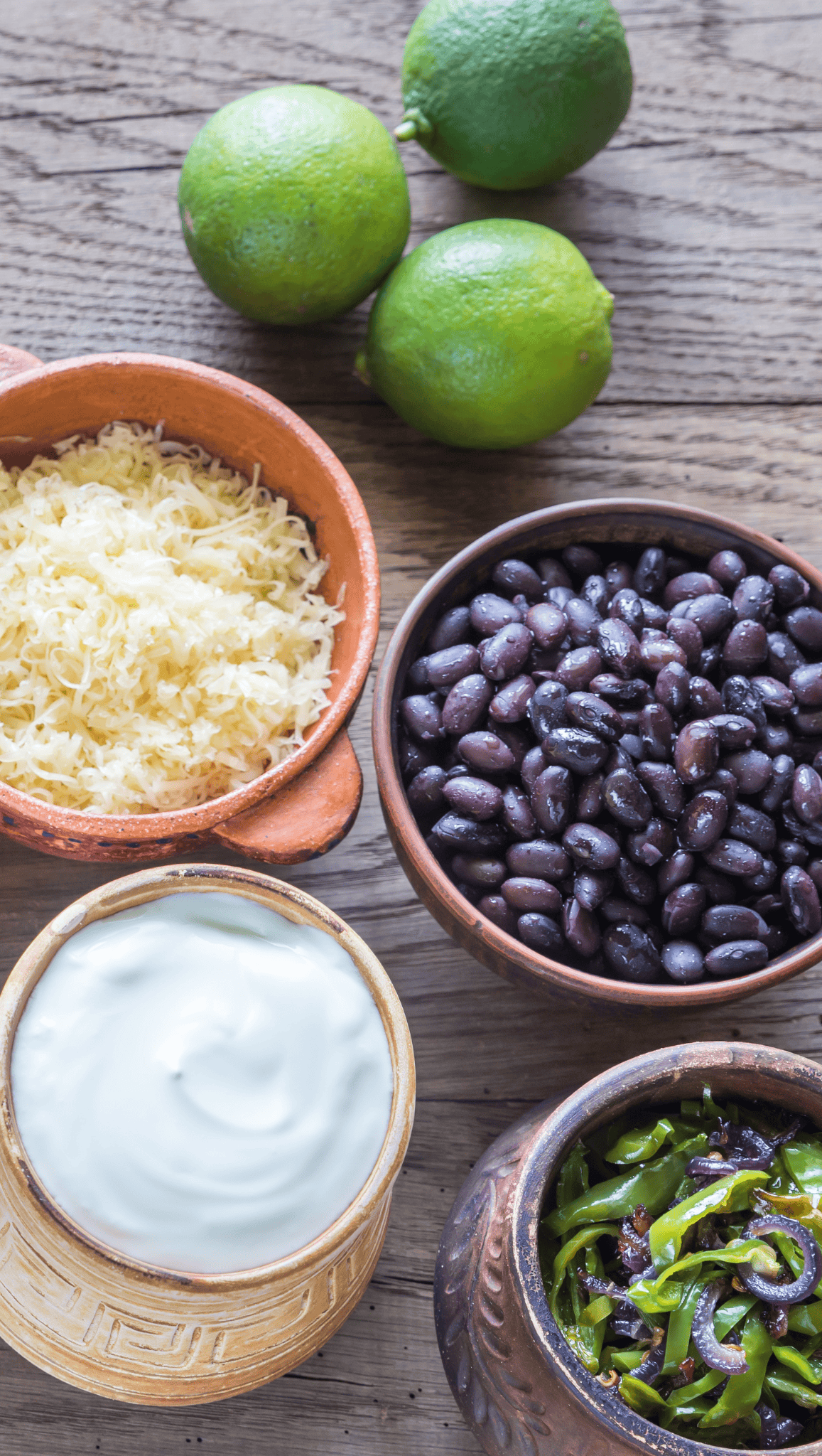 Four bowls on a wooden table display shredded cheese, black beans, sour cream, and sautéed green peppers—perfect toppings for a vibrant Mexican Salad Bowl. Three whole limes rest in the background, adding a fresh citrus touch.