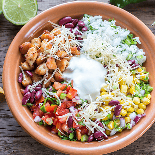 A colorful Mexican Salad Bowl with grilled chicken, rice, beans, corn, cheese, salsa, and sour cream, surrounded by tortilla chips, lime halves, and green chili peppers on a wooden table.