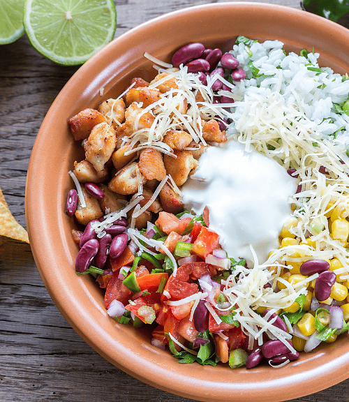 A colorful Mexican Salad Bowl with grilled chicken, rice, beans, corn, cheese, salsa, and sour cream, surrounded by tortilla chips, lime halves, and green chili peppers on a wooden table.
