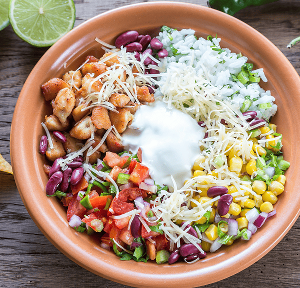 A colorful Mexican Salad Bowl with grilled chicken, rice, beans, corn, cheese, salsa, and sour cream, surrounded by tortilla chips, lime halves, and green chili peppers on a wooden table.