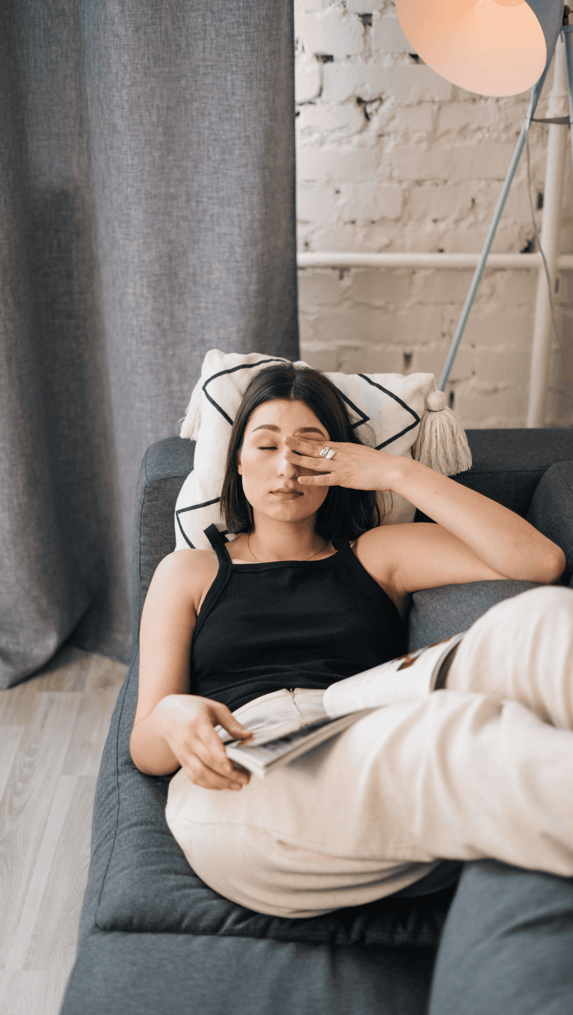 A woman in casual clothes sits on a couch, holding a book in one hand and rubbing her eyes with the other, looking tired or stressed—perhaps her nervous system is feeling overwhelmed. A lamp and gray curtains are in the background.
