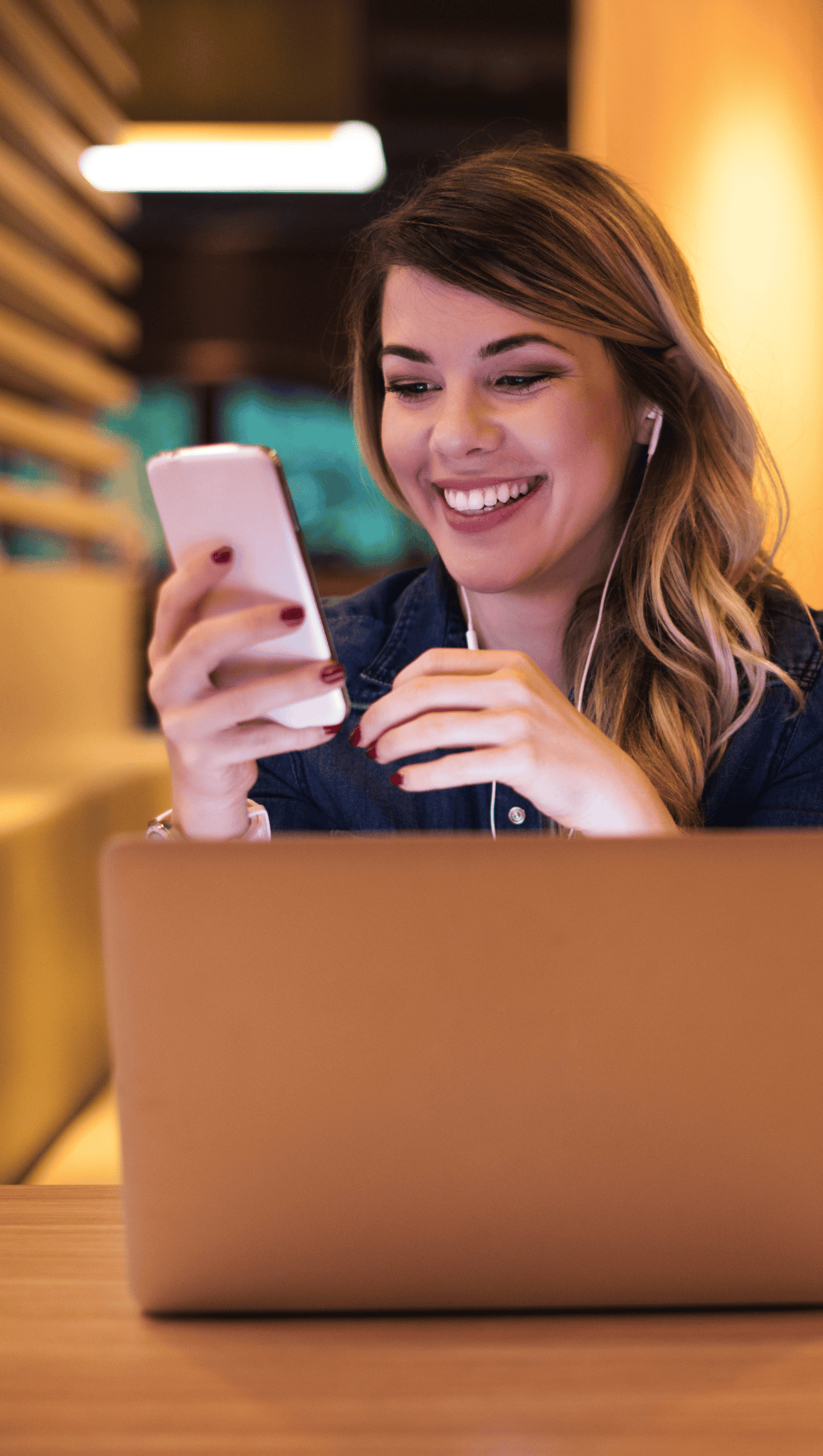 A smiling woman with long hair sits at a table with a laptop, holding a smartphone and wearing earphones, appearing engaged and happy in a warmly lit indoor setting, as she learns about the nervous system.