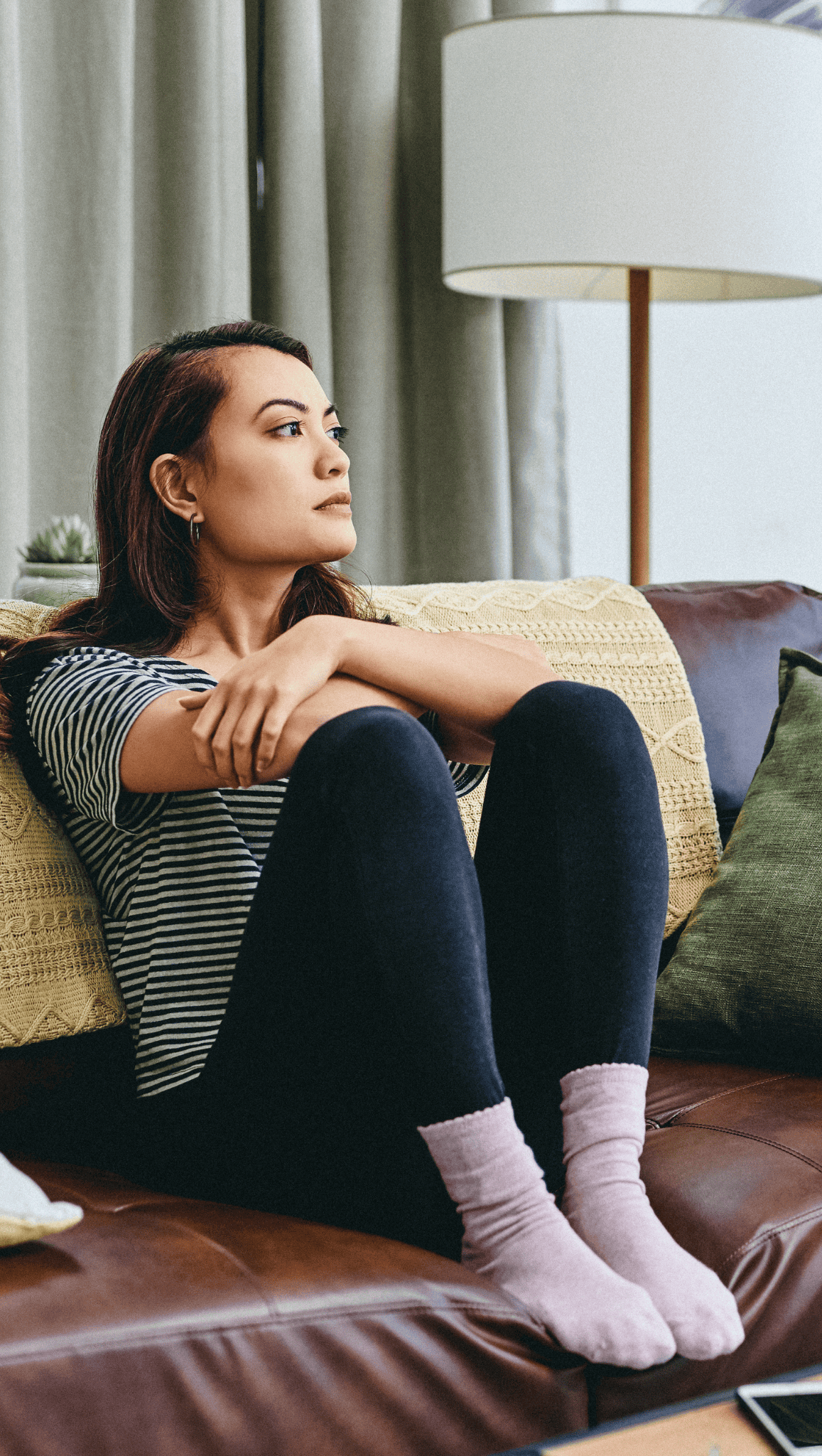 A woman with brown hair sits on a couch, hugging her knees and looking thoughtfully out of frame. She wears a striped shirt, black leggings, and light pink socks, as if deep in thought about her nervous system. A lamp and pillows are in the background.