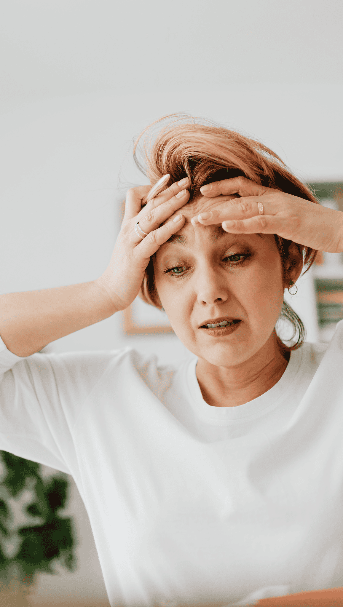 A woman in a white shirt holds her forehead with both hands and looks worried or stressed, her tense expression hinting at the strain on her nervous system.