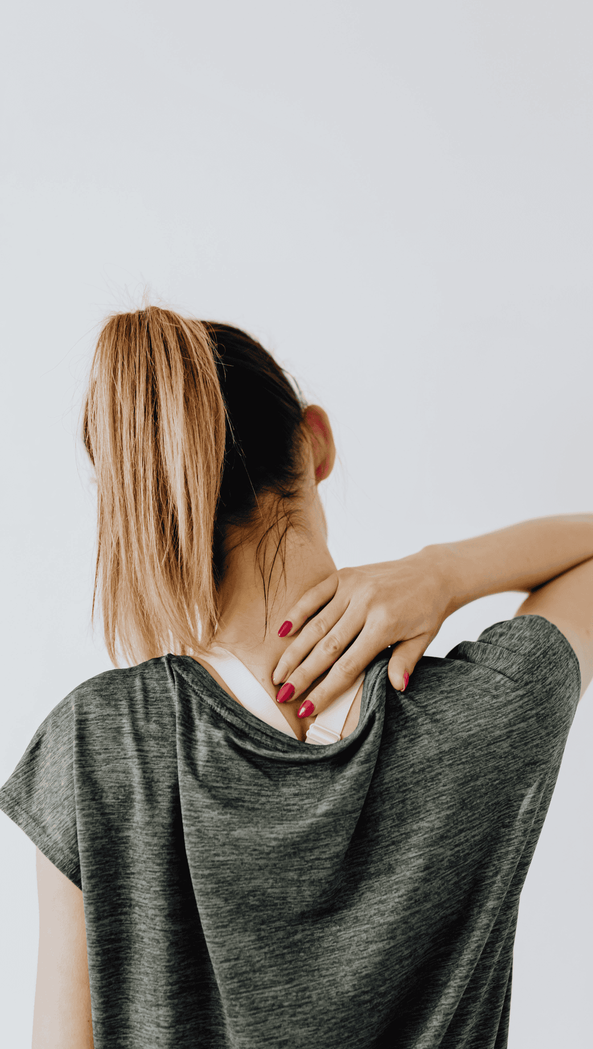 A woman with a ponytail, seen from behind, touches the back of her neck with one hand, suggesting discomfort in her nervous system. She wears a grey t-shirt and has red painted nails.