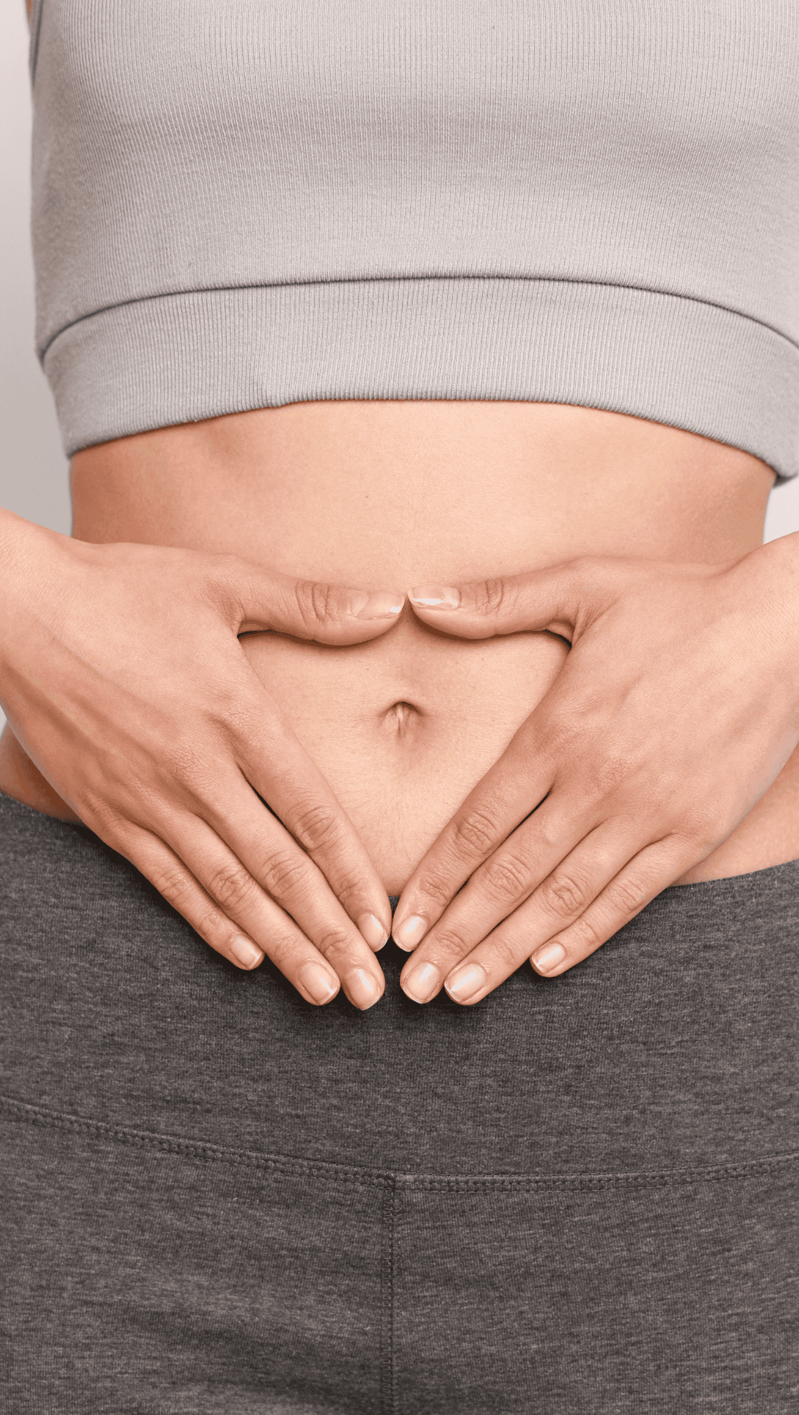 Close-up of a person in a gray crop top and pants, hands forming a heart shape over their bare stomach, centered around the navel—a subtle nod to the vital connection between gut health and the nervous system.