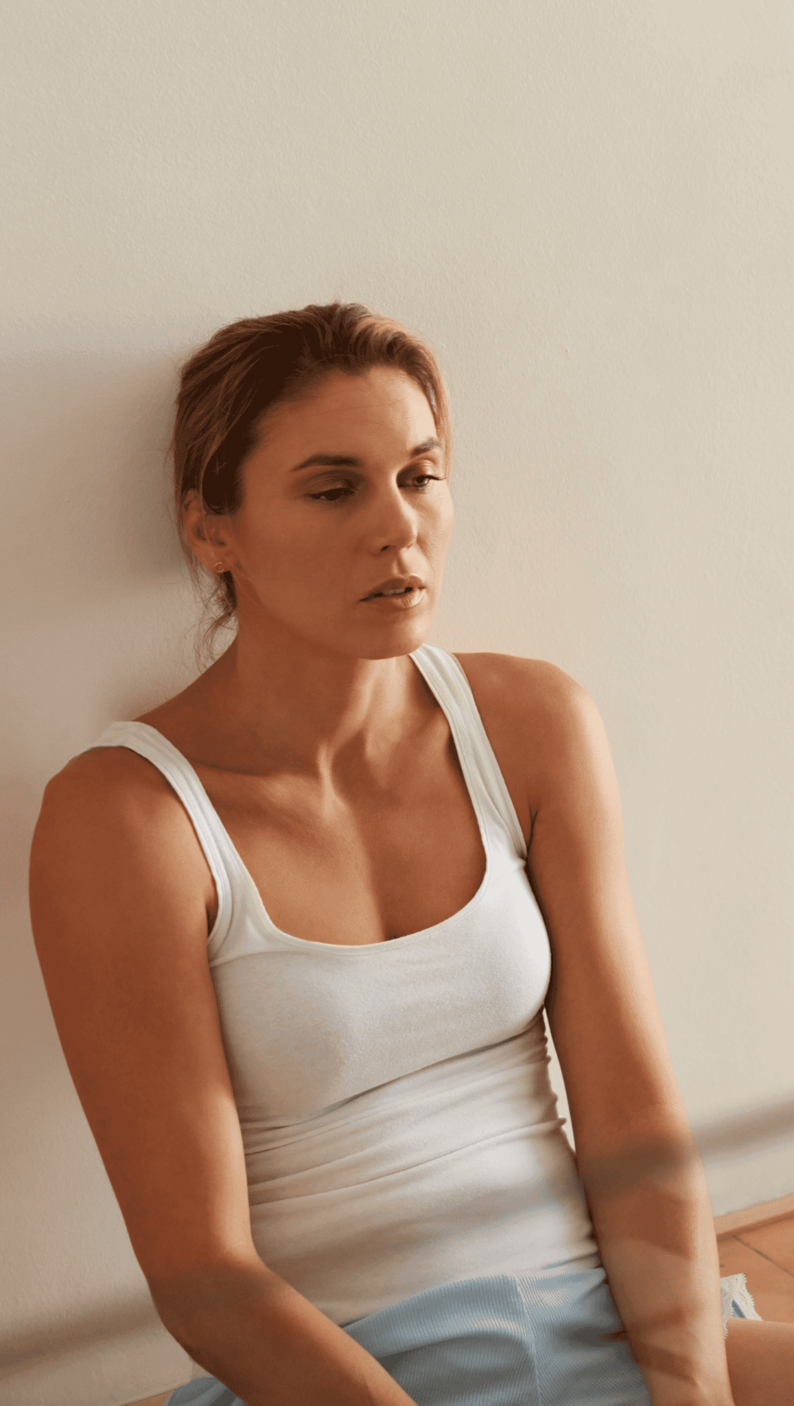 A woman with light brown hair wearing a white tank top sits against a light-colored wall, looking tired or pensive—her expression perhaps reflecting the strain on her nervous system—as her eyes half-close and her arms rest at her sides.