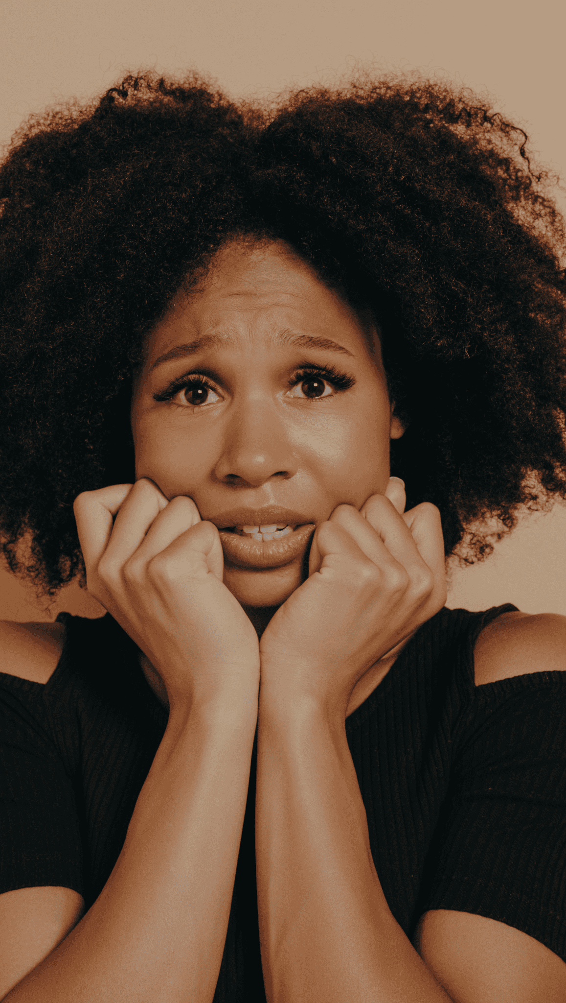 A woman with curly hair and a black top holds her hands to her face, looking anxious or worried, her wide eyes and furrowed brows revealing the strain on her nervous system, against a plain background.