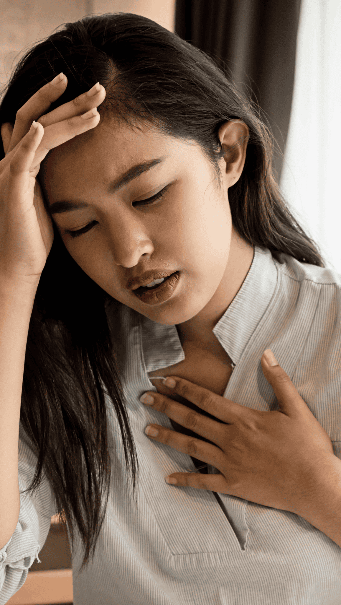 A young woman with long dark hair touches her forehead and chest, eyes closed and expression pained, possibly feeling unwell or stressed. She may be experiencing discomfort related to the nervous system while indoors in a light-colored top.