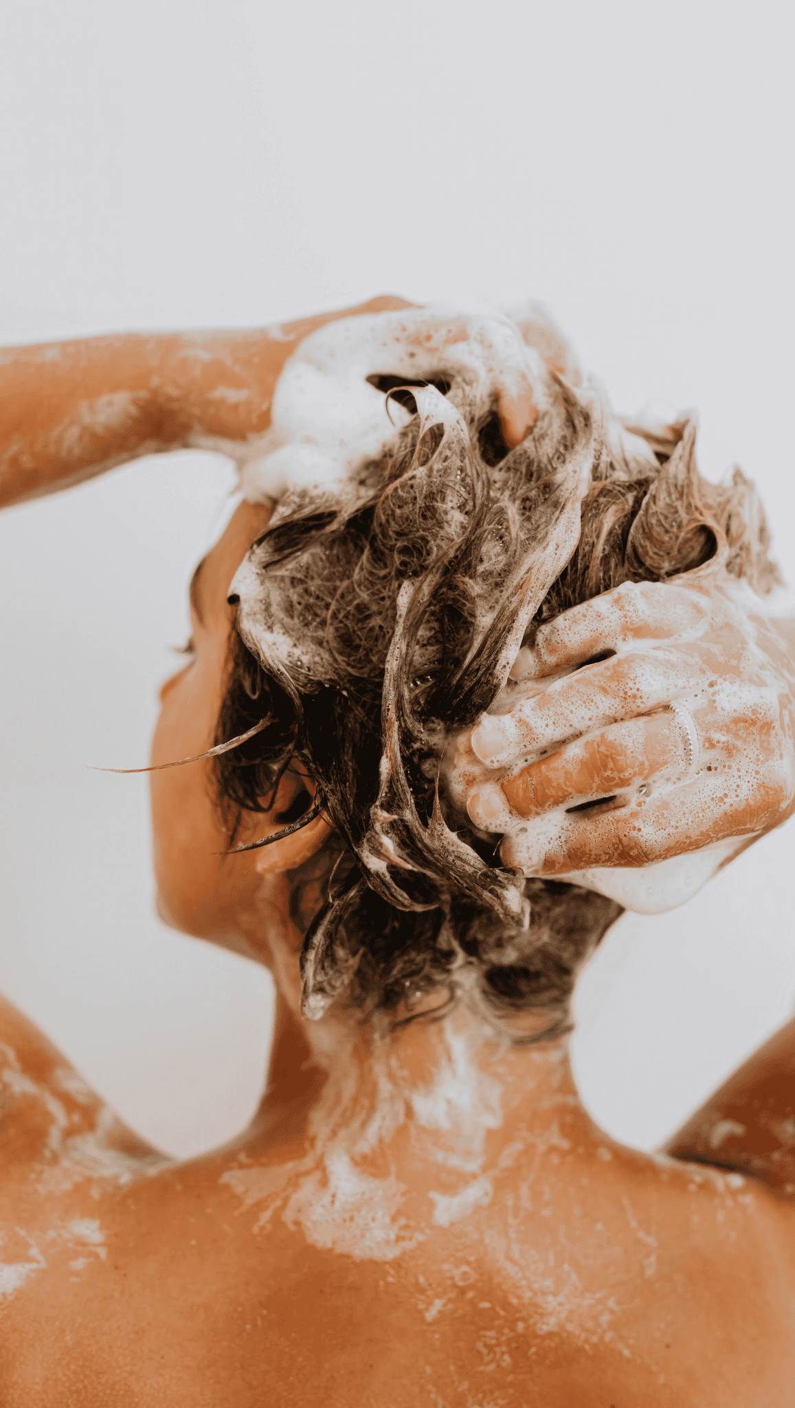 A person with short, dark hair is seen from behind, washing their hair with shampoo during their Night Shower Routine, creating lots of white, foamy lather as they massage their scalp. The background is plain white.