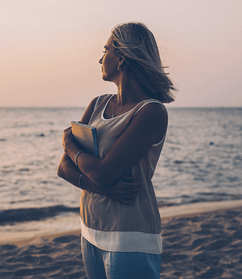A woman stands on a sandy beach at sunset, holding a book to her chest and looking out at the calm sea—lost in thought, she embraces the peaceful atmosphere and contemplates mindset shifts beneath the pastel sky.