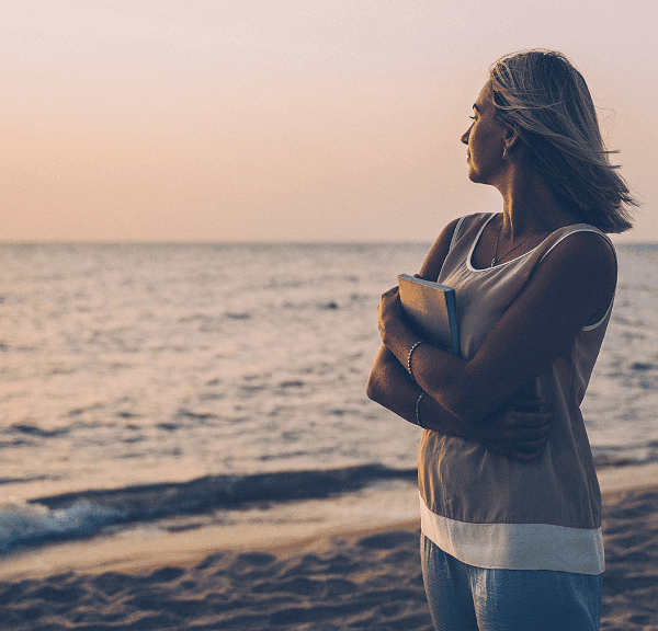 A woman stands on a sandy beach at sunset, holding a book to her chest and looking out at the calm sea—lost in thought, she embraces the peaceful atmosphere and contemplates mindset shifts beneath the pastel sky.