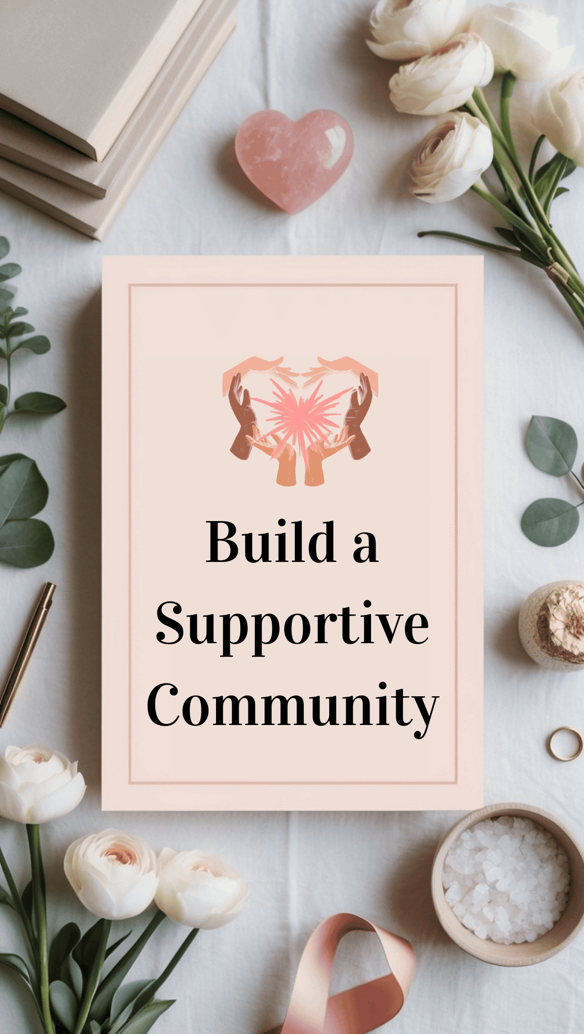 A notebook with Build a Supportive Community on the cover, surrounded by white flowers, a pink heart crystal, books, a ribbon, and a bowl of bath salts on a white cloth background—perfect for self-care and nurturing a positive mindset.