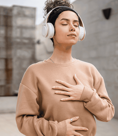 A woman wearing headphones and a beige sweatshirt stands outdoors with eyes closed, one hand on her chest and the other on her stomach, appearing calm as she takes a moment to self-soothe.
