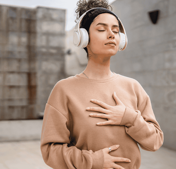 A woman wearing headphones and a beige sweatshirt stands outdoors with eyes closed, one hand on her chest and the other on her stomach, appearing calm as she takes a moment to self-soothe.